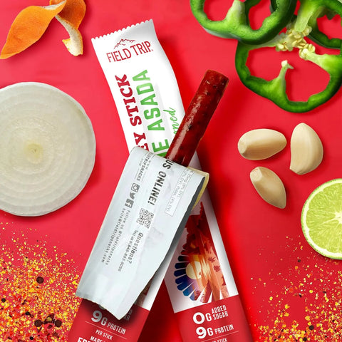 A protein-rich snack, the Field Trip Carne Asada Turkey Stick 0.5oz sits on a red surface with its wrapper partly removed, surrounded by garlic cloves, green pepper slices, onion, lime, dried chili flakes, and orange peel.