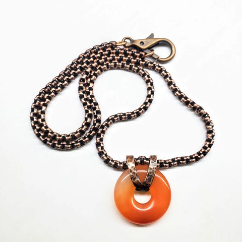 The Carnelian Gemstone Donut Copper Chain Necklace by Alexa Martha Designs features a round orange carnelian pendant with a metal loop, handcrafted for rustic style jewelry lovers.