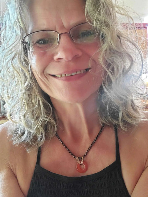 A woman with wavy, shoulder-length blonde hair and glasses smiles at the camera, wearing a black top and the Alexa Martha Designs Carnelian Gemstone Donut Copper Chain Necklace.