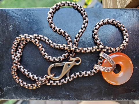 The Carnelian Gemstone Donut Copper Chain Necklace by Alexa Martha Designs features a handcrafted copper pendant with a round orange carnelian gemstone on a bronze-colored chain, secured by a lobster clasp and displayed on a dark surface.