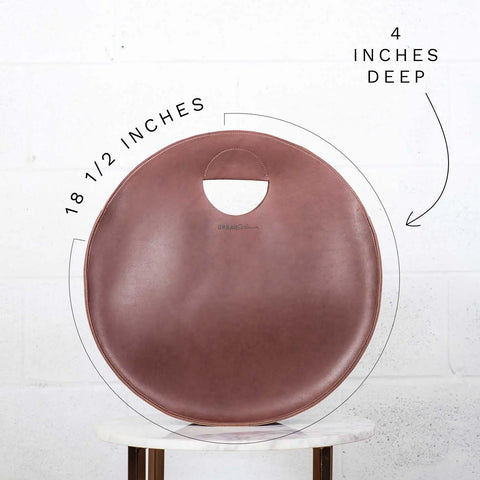 The Urban Southern Carrollton Circle Tote, a round full-grain leather bag with a circular handle (18.5 wide, 4 deep), sits on a small table against a white brick wall.