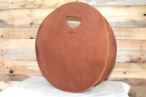 The Urban Southern Carrollton Circle Tote, crafted from full-grain brown leather with visible stitching and a cutout handle, stands upright on a white surface against a wooden plank background. Its round shape features a simple, minimalist design.