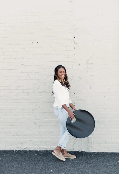 A woman in a white blouse and light blue jeans smiles in front of a pale brick wall, holding a round black reflector and carrying the Urban Southern Carrollton Circle Tote made from full-grain leather.