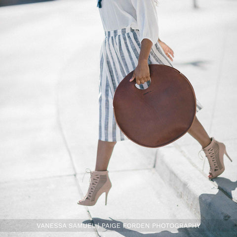 A woman in a white blouse, striped skirt, and high-heeled lace-up sandals carries the Carrollton Circle Tote by Urban Southern as she steps off a curb in bright sunlight.