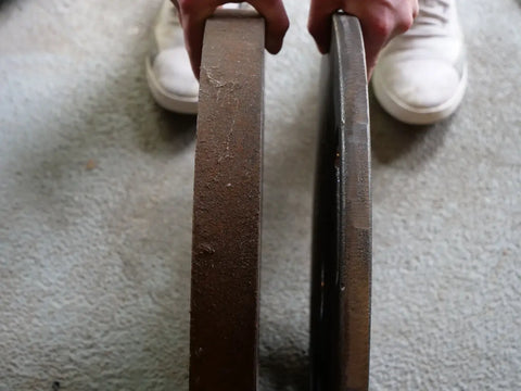 Close-up of two Weight It Out Cast Iron Machined Weight Plate Pairs held upright by hands over a carpeted floor, with white shoes visible in the background.