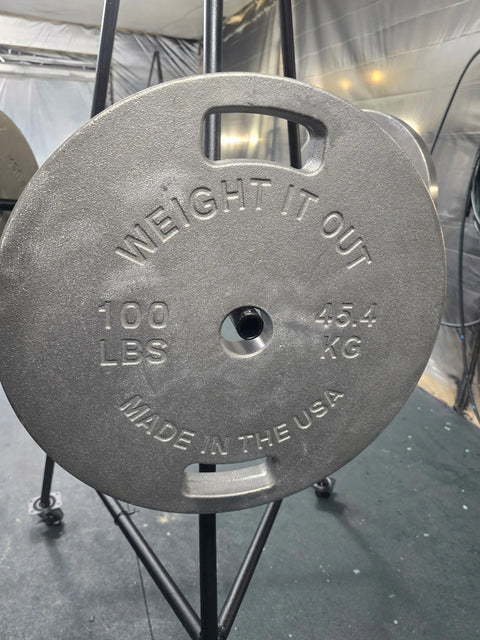 A close-up of a 100-pound Cast Iron Machined Weight Plate Pair by Weight It Out, labeled MADE IN THE USA, mounted on a black metal rack in a gym setting.