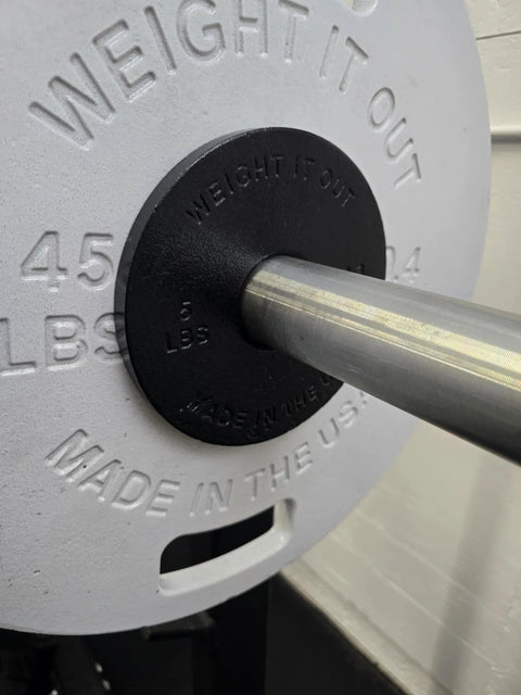 Close-up of a barbell with a large white Weight It Out Cast Iron Weight Plate Set and a black 5 lb collar on the bar. Ideal for anyone looking for durable, USA-made cast iron plates for their workouts.