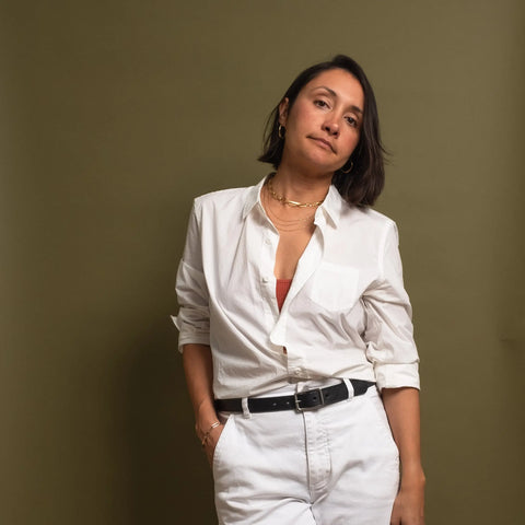 A person with short dark hair poses confidently in a white button-up shirt, white pants, and gold jewelry, highlighted by the CASUPO Smith Thinner Belt Black with a solid brass buckle, against an olive-green background.