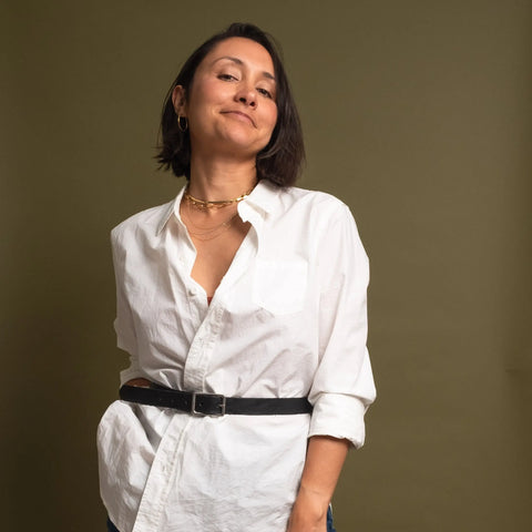 A person with short dark hair, wearing a white button-up shirt cinched with the CASUPO Smith Thinner Belt Black and gold jewelry, stands confidently against an olive green background, looking slightly upwards and smiling softly.