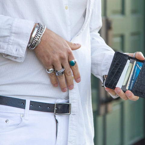 A person wearing a white shirt and pants holds an open black wallet, accessorized with silver rings, bracelets, and a green stone ring. The look is finished with the CASUPO Smith Thinner Belt Black with solid brass buckle. A blurred door appears in the background.