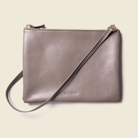 The CASUPO Crossbody Bag - Misty Lilac features sustainable leather in a soft hue, a slim adjustable strap, minimal design, gold-tone zipper, and subtle embossed branding near the bottom.