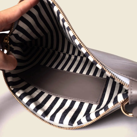 A hand holds open the CASUPO Crossbody Bag - Misty Lilac, showing a black and white striped lining and a rectangular pocket made from sustainable leather inside.