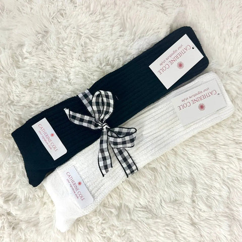 The Catherine Cole 2 Pairs Bundle ULTIMATE Slouch Socks in black and white, accented with a plaid ribbon, evoke retro 80s and 90s vintage style. Both pairs feature ribbed texture and branded tags, displayed on a soft white surface.