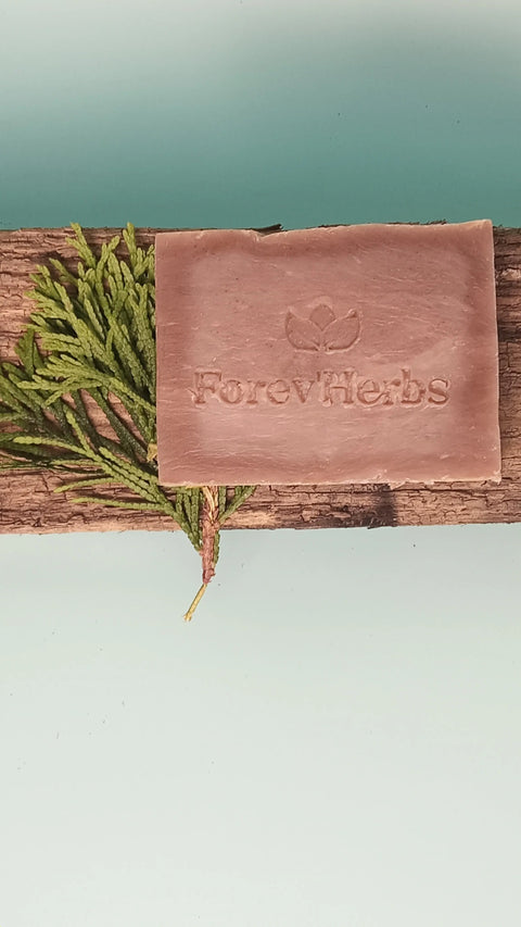 A rectangular Cedarwood Sage Handmade Soap by Forev'Herbs, engraved with the brand name, rests on a rustic wooden plank with green fir leaves, set against a pale blue background.