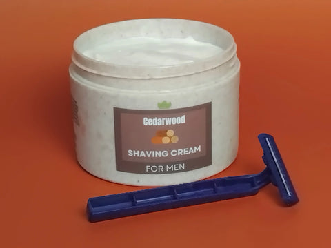 A jar of Forev'Herbs Cedarwood Shaving Cream sits on an orange surface beside a blue disposable razor, perfect for helping reduce razor bumps.