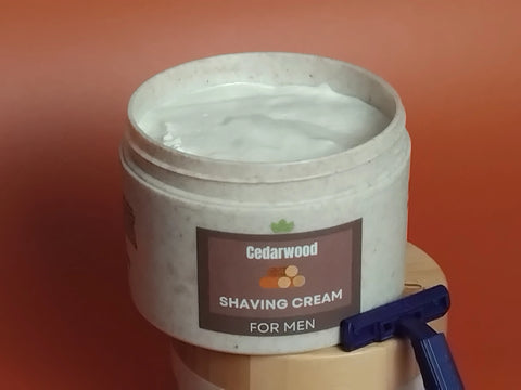 Forev'Herbs Cedarwood Shaving Cream, infused with tea tree oil, sits open on a wooden surface with a blue razor beside it. The solid orange background highlights this blend made to help eliminate razor bumps.