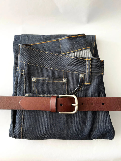 A pair of dark blue jeans folded with the Rogue Industries Chamberlain Leather Belt (1.25 wide) threaded through the loops, displayed on a white background.
