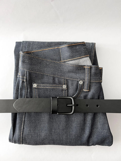A pair of dark blue denim jeans is neatly folded with the Rogue Industries Chamberlain Leather Belt (1.25 wide) threaded through the belt loops, all displayed on a plain white background.