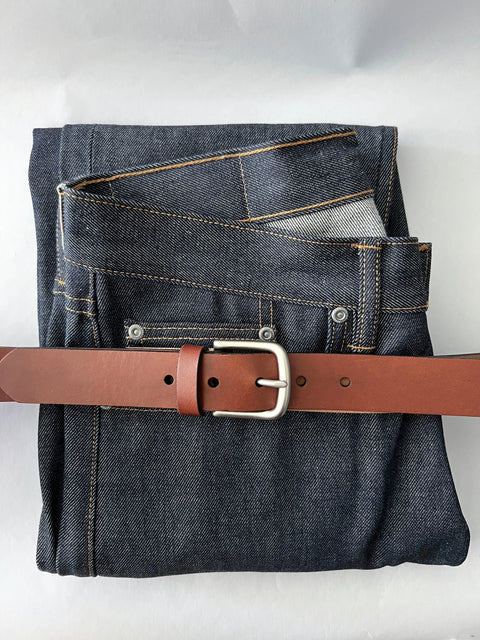 A pair of dark blue jeans neatly folded with the Rogue Industries Chamberlain Leather Belt (1.25 wide) threaded through the belt loops, displayed on a light background.