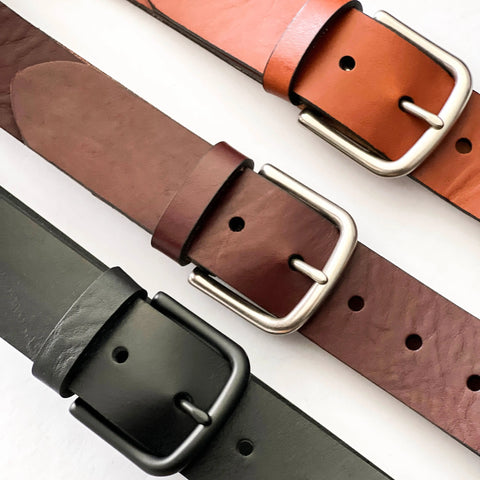 Three Chamberlain Leather Belts by Rogue Industries—tan, brown, and black—are arranged diagonally on a white backdrop. Each 1.25-wide handcrafted belt features a rectangular metal buckle in different finishes.