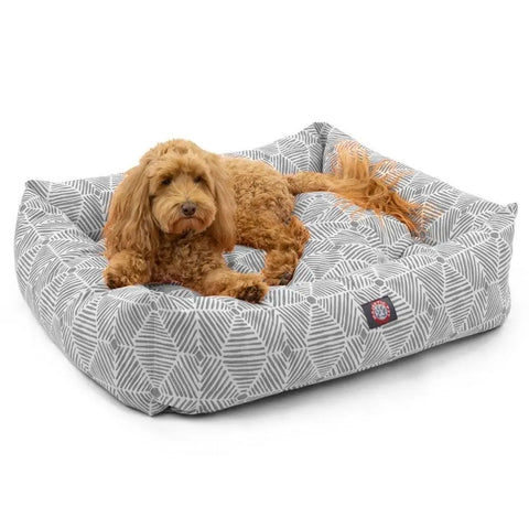 A curly-haired brown dog is lying on the Majestic Pet Charlie Luxe Bagel Bed, a gray and white geometric-patterned, machine washable bed with raised edges.