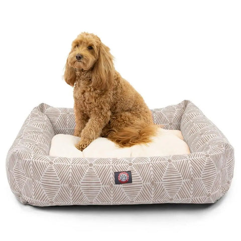 A curly-haired, light brown dog sits on the Majestic Pet Charlie Luxe Bagel Bed, a beige and white patterned bed with raised edges, looking slightly to the side.