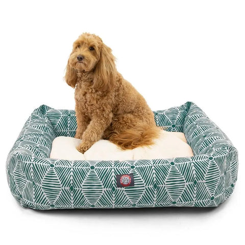 A fluffy, light brown dog sits on the Majestic Pet Charlie Luxe Bagel Bed, a cushioned rectangular bed with a green and white geometric pattern made from durable upholstery-grade fabrics, set against a white background.