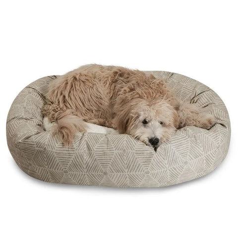 A fluffy, light brown dog is resting on the Charlie Sherpa Bagel Dog Bed by Majestic Pet, which features a round shape and soft cotton twill with a beige geometric pattern for comfort.