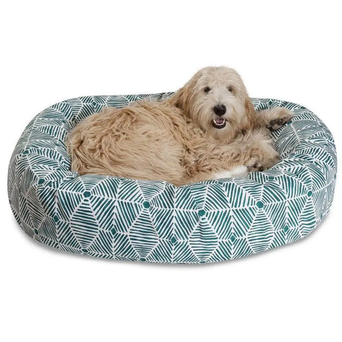 A fluffy, light brown dog is relaxing in a Majestic Pet Charlie Sherpa Bagel Dog Bed featuring a blue and white geometric pattern made of soft cotton twill. The dogs mouth is open, looking relaxed and content.