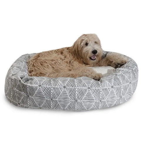 A fluffy, light brown dog relaxes on the Majestic Pet Charlie Sherpa Bagel Dog Bed, a round cotton twill bed with a gray and white pattern, looking at the camera with its mouth slightly open.