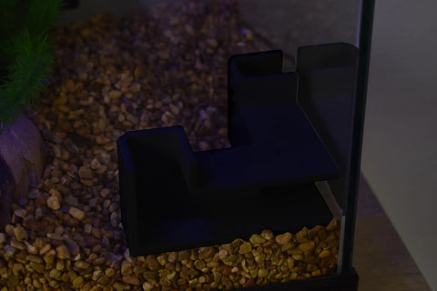 The Chatelet Aquarium Underground Tunnel by Chatelet Manufacturing sits on brown pebbles beside the glass, with green plant leaves and driftwood partially visible, adding a unique touch to your aquarium decor.