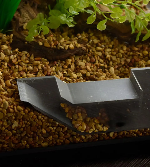 A close-up of an aquarium corner displays brown gravel, a plastic plant, driftwood, and the Chatelet Aquarium Underground Tunnel by Chatelet Manufacturing, a unique gray hideout partially buried for fish and shrimp. Made in USA.