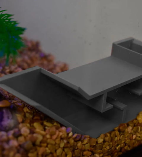 The Chatelet Aquarium Underground Tunnel by Chatelet Manufacturing sits on brown gravel, paired with a green plastic plant and blurred background, highlighting this unique fish and shrimp hideout for aquariums.