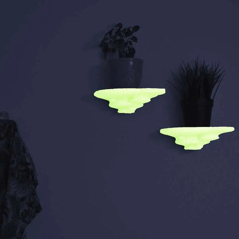 Two small potted plants sit on Chatelet Mushroom Floating Shelves by Chatelet Manufacturing, their scalloped, wall-mounted design glowing against a dark background as a patterned cloth peeks in from the left.