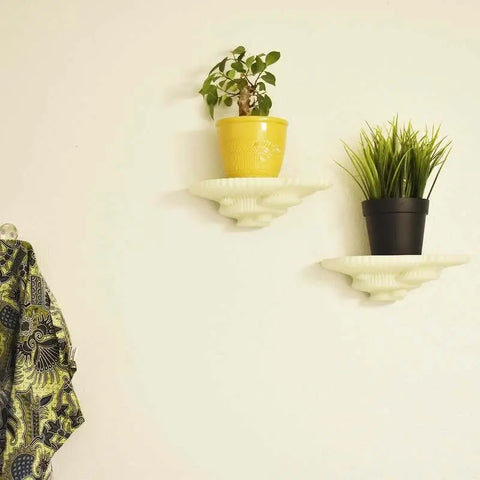 Two Chatelet Mushroom Floating Shelves by Chatelet Manufacturing each display a potted plant—one in a yellow pot with leafy foliage, one black with grass-like leaves. Nearby, a patterned fabric on a hook accents your light-colored wall décor.