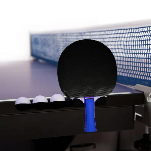 A Chatelet Ping Pong Table Caddy (Pack of 2) by Chatelet Manufacturing rests on the table’s edge next to four white balls, a black paddle with a blue handle, and the net visible in the background.