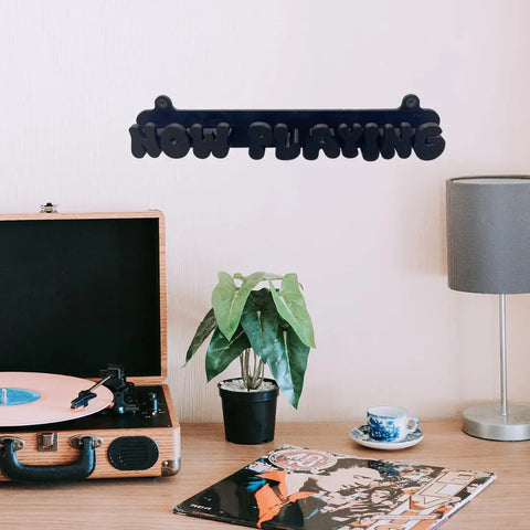 A tabletop with a record player spinning peach vinyl, a plant, blue teacup and lamp sits below the Chatelet Manufacturing Groovy Now Playing Vinyl Record Wall Mount—a retro display sign made in the USA.