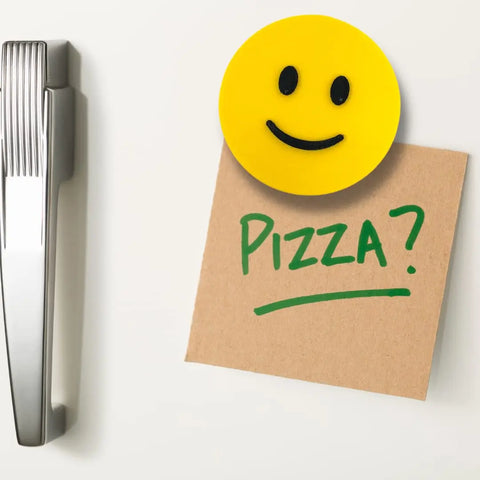 A black and yellow Emoji Smiley Face from Chatelet Manufacturing holds a brown note with Pizza? in green marker on a white fridge near a metallic handle—great as a fun gift. Made in USA.