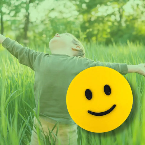 A joyful child stands in tall grass with outstretched arms, while a large black and yellow Chatelet Manufacturing Emoji Smiley Face Decorative Standalone Ideogram appears in the green-tinted foreground. Made in USA.