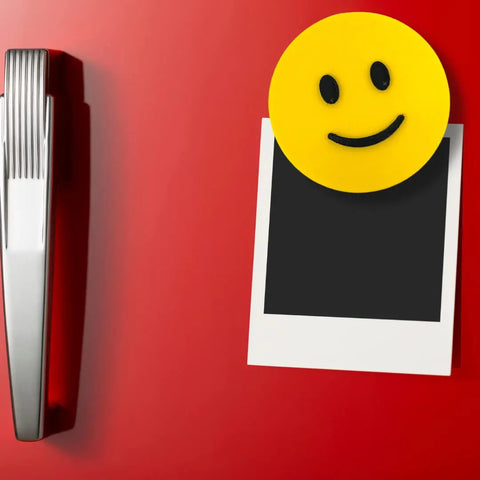 A silver stapler and a blank instant photo with a Chatelet Manufacturing Emoji Smiley Face Decorative Standalone Ideogram (black and yellow, made in USA) sit on a red surface.