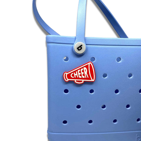 A light blue Bogg Bag features a Chatelet Manufacturing BOGLETS red megaphone cheerleading charm, attached through the small holes on the front. The handle is at the top for easy carrying.