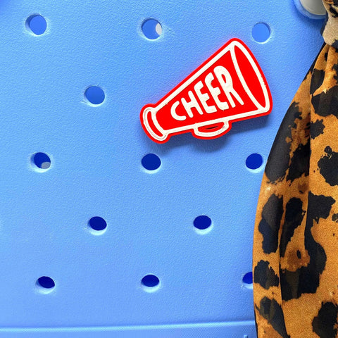 A red megaphone BOGLETS Cheerleading Charm by Chatelet Manufacturing, featuring CHEER in white, decorates a light blue Bogg Bag; a leopard print fabric is draped on the right side.