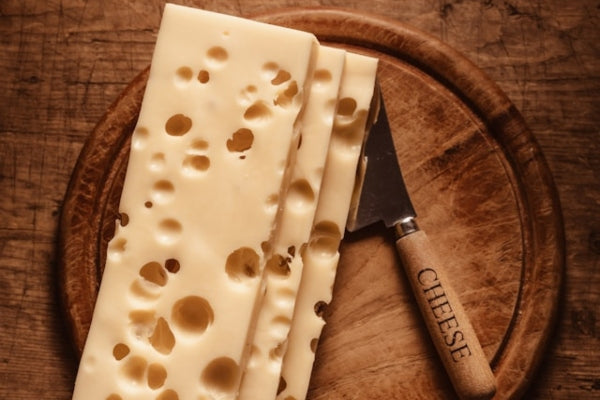 Two slices of Swiss cheese on a wooden cutting board with a knife.