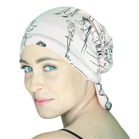 A light-skinned woman with blue eyes models the Chemo Beanies® Stephanie beanie in light pink with black designs. She looks over her shoulder, softly smiling, against a plain white background.