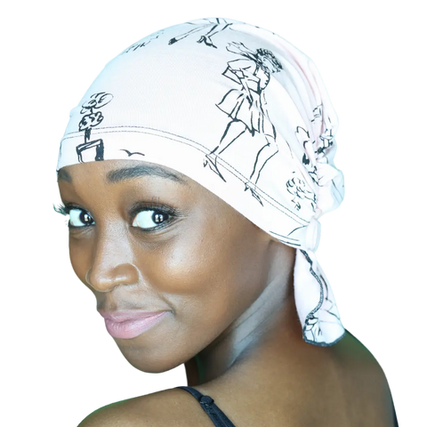 A woman with smooth dark skin wears the Chemo Beanies® Stephanie, a light-colored chemo scarf with fashion sketch designs, and a black strap top. She smiles gently over her shoulder at the camera against a plain background.