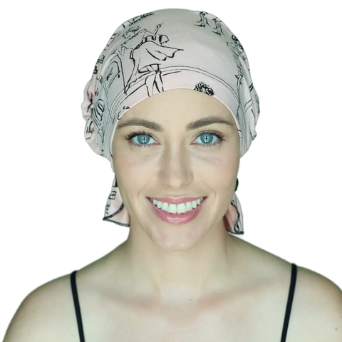 A smiling woman with bright blue eyes wears the Chemo Beanies® Stephanie scarf in light pink with a black pattern, paired with a black spaghetti strap top, against a plain background.