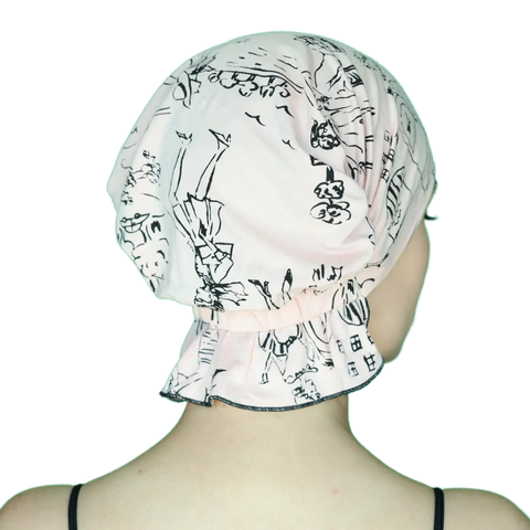 A person is seen from behind wearing the Chemo Beanies® Stephanie slip-on beanie in light pink with black illustrations, fully covering their hair and head—ideal cancer headwear. Thin black top straps are visible on their shoulders.
