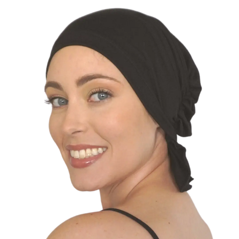 A smiling woman with blue eyes and light skin wears the Chemo Beanies® Margaret black cancer headwear scarf and a black sleeveless top, looking over her shoulder against a plain white background.