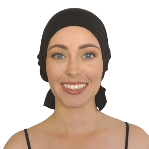 A woman with light skin, blue eyes, and natural makeup smiles at the camera on a plain background, wearing the Margaret Chemo Beanie® by Chemo Beanies® and a black strap top.