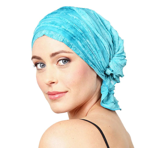 A woman with light skin and blue eyes wears the Chemo Beanies® Molly chemo beanie in bright blue and a black spaghetti strap top, looking over her shoulder with a gentle smile against a white background.